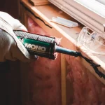 Gloved hand caulking a crack beside a window